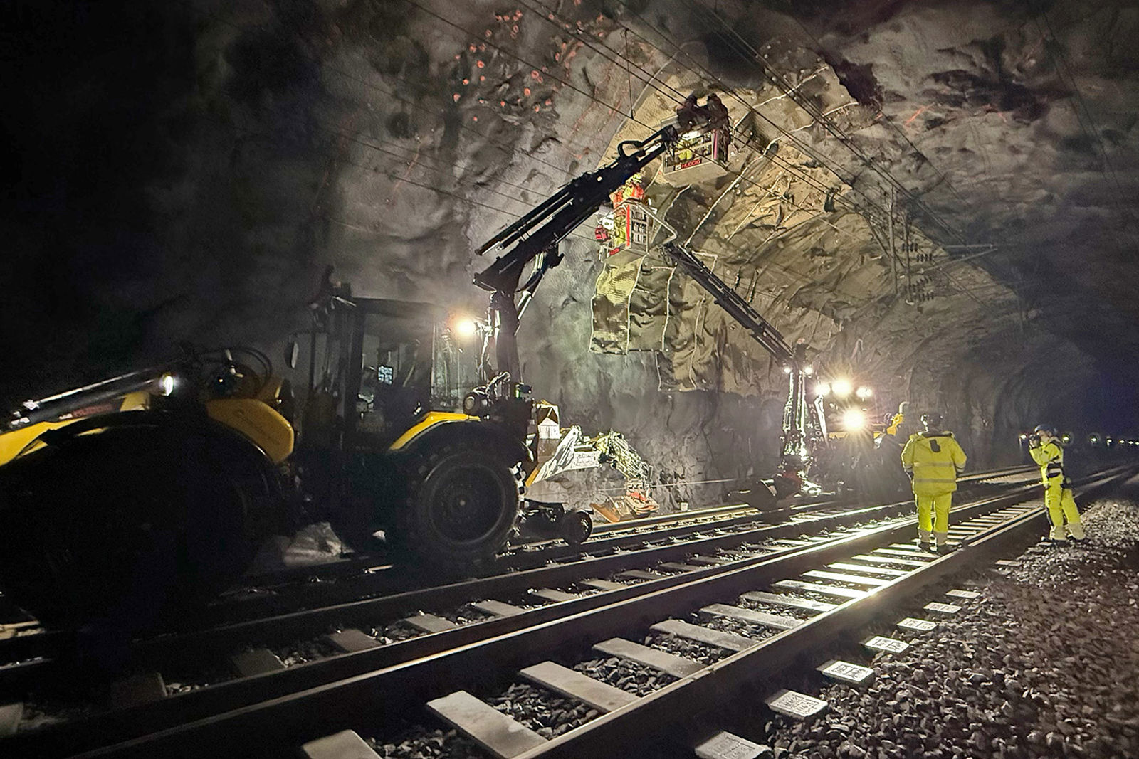 To skinne/vei-maskiner med lifter hvor mannskaper jobber med montering av matter i tunneltaket.