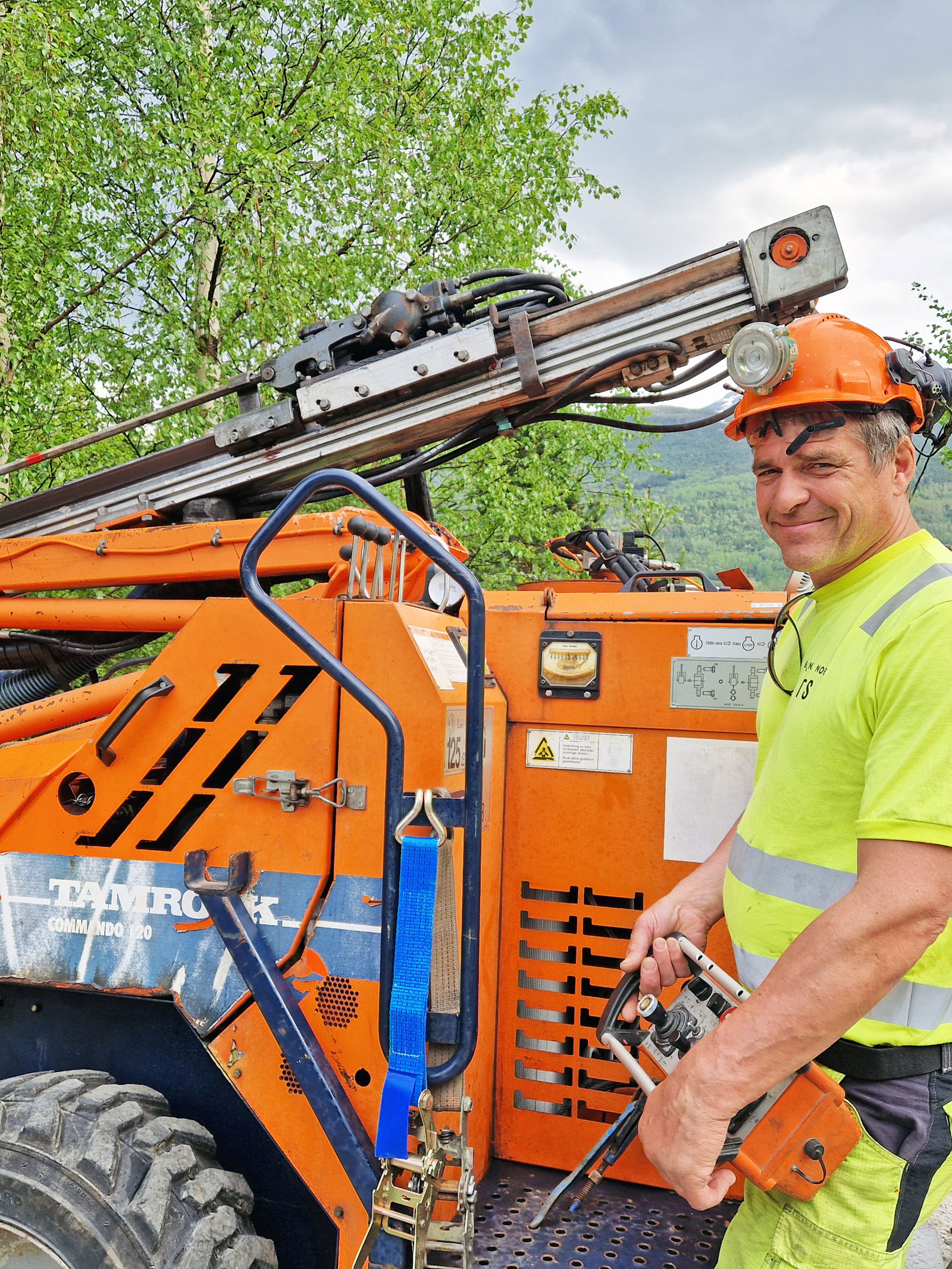 En mann i vernetøy står foran en oransje anleggsmaskin, som er en borerigg. 