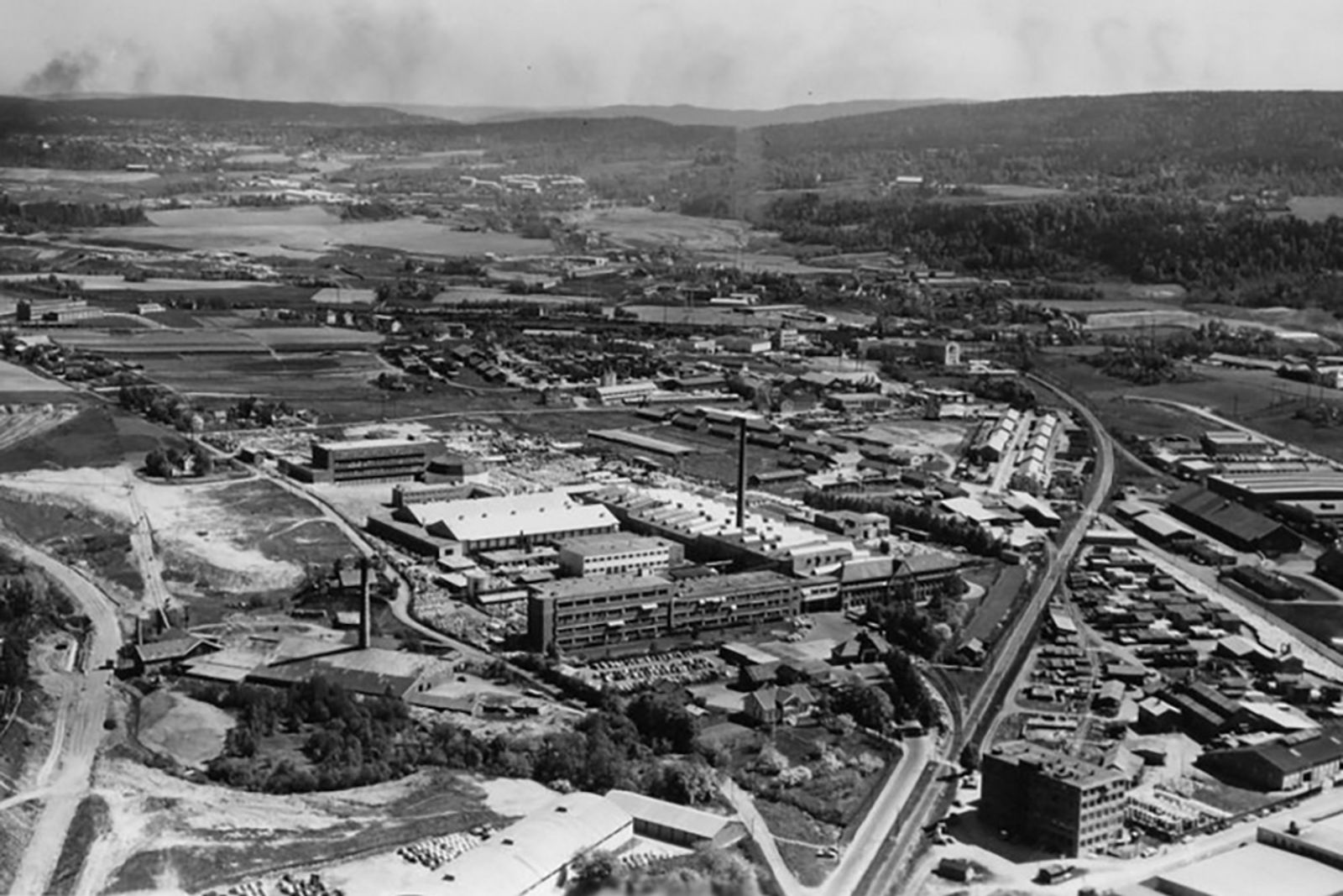 Stort fabrikkområde omgitt av bane, veier og bebyggelse. Jorder i bakgrunnen.