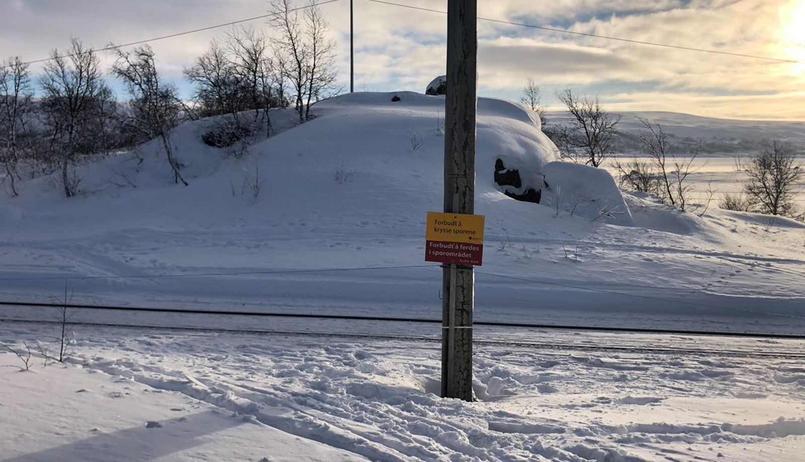 I et snødekt terreng står det en stolpe med advarsel om å krysse jernbanen. Ved siden av går skispor som krysser jernbanen.