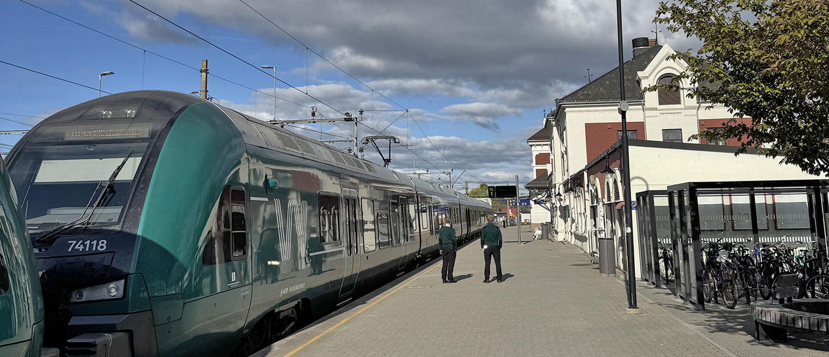 Grønt Vy-tog til venstre, personer på plattformen, stasjonsbygningen og sykkelparkering til høyre.
