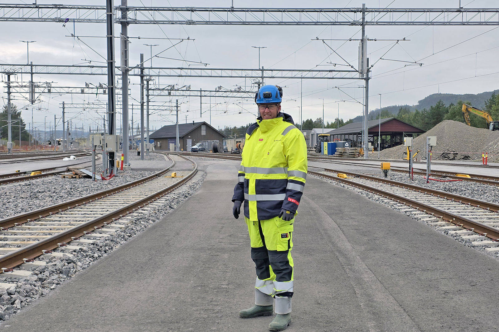 Prosjektlederen iført vernetøy og hjelm med spor og bygninger i bakgrunnen.