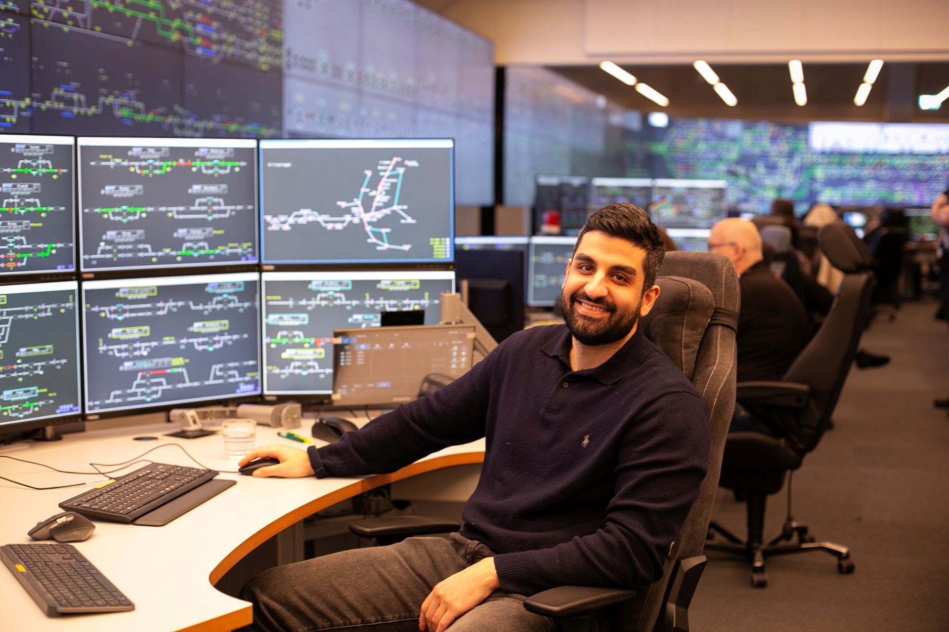 Azad sitter foran flere dataskjermer med systemer for en togleder og smiler til kamera